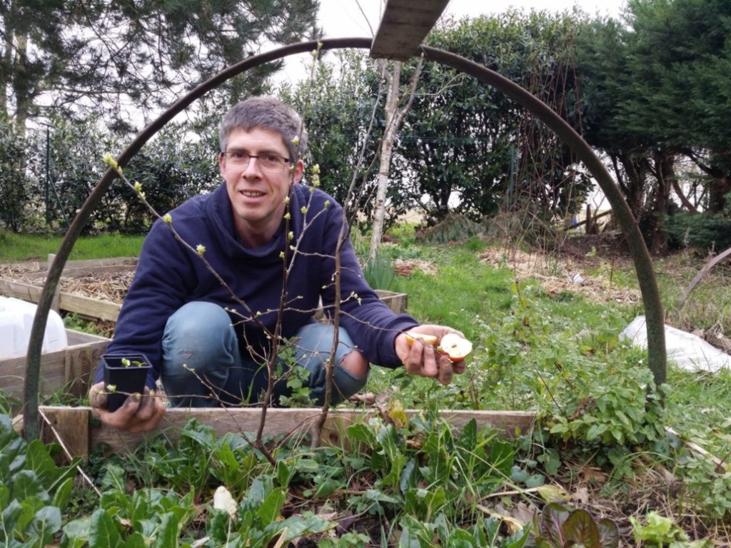 Bertrand souriant accroupi devant une planche végétale de son jardin, montre deux demi-pommes et un petit pot avec un plant. L’image le montre comme entouré par un arceau en demi-cercle métallique probablement support d’une petite serre-tunnel.